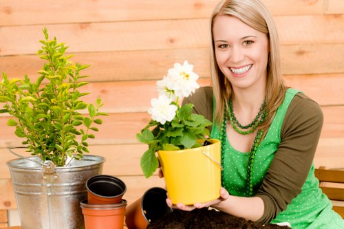 The image shows a cheerful woman with long blonde hair, smiling as she holds a yellow ceramic pot with white flowering plants, possibly indoor garden or patio plants. She is dressed casually in a green polka-dot top layered with a brown cardigan, and wears a beaded green necklace. In front of her on a wooden table are various gardening supplies, including an empty small terracotta pot, a black plastic pot, and a metal bucket containing a small, bright green shrub with dense foliage. The background features a wooden fence or wall, suggesting an outdoor or patio setting, with natural daylight illuminating the scene. The overall environment appears to be a garden or outdoor space dedicated to gardening activities, with a focus on plant care and nurturing, fitting within the context of gardening services offered by Lawn Mowing Archway in the local area around London or North London postcodes.